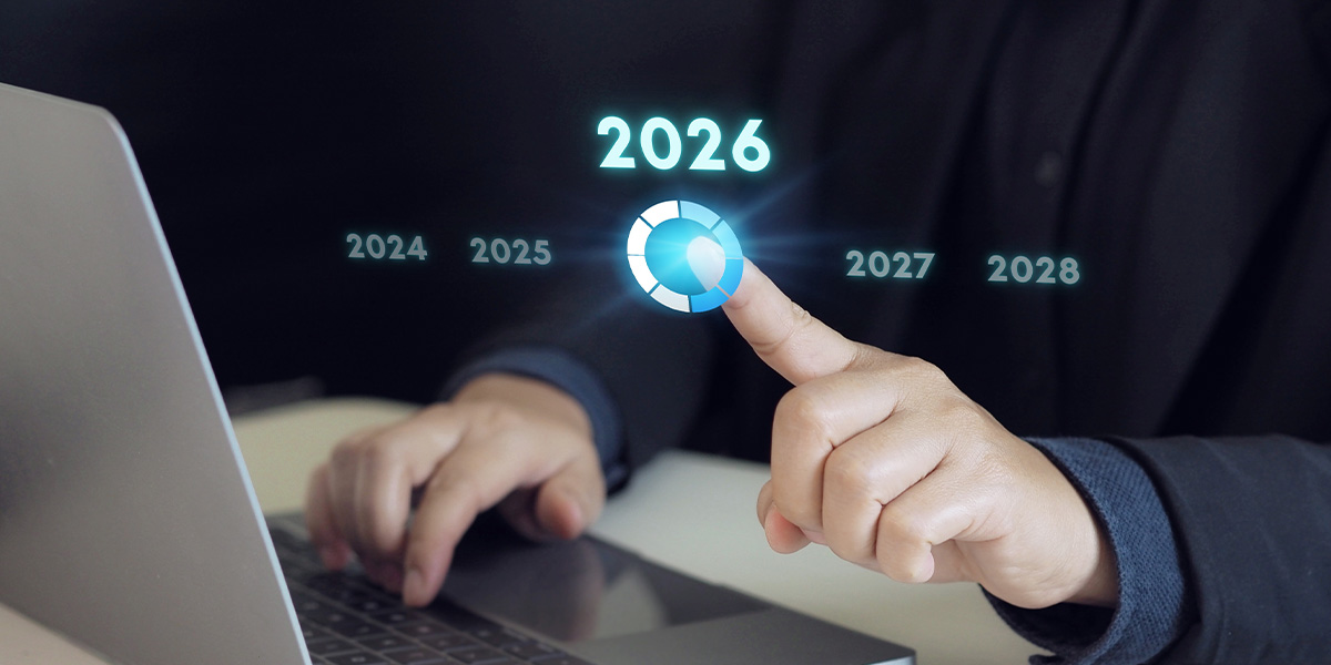 Person in a dark suit seated at a desk using a laptop, pointing at a glowing circular graphic labeled “2026.” Surrounding years 2024, 2025, 2027, and 2028 appear in smaller text against a dark background.